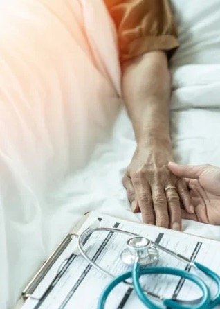 Caregiver gently holding the hand of a resident resting in bed, beside a clipboard and stethoscope